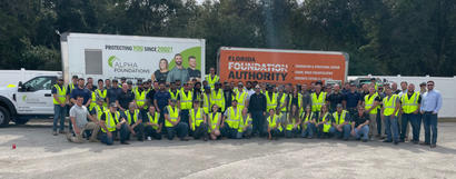 A large group of people in safety vests posing in front of two trucks with company logos.