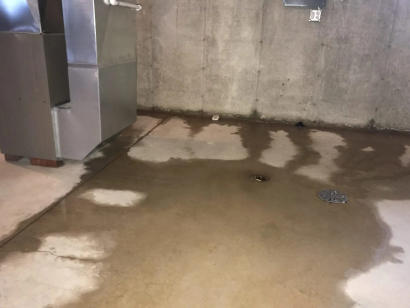 Basement floor with water pooling around a drain near a furnace against a bare concrete wall.