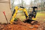 A person operates a yellow mini excavator, digging soil near a house in a wooded area. A red shovel is nearby.
