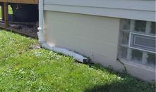 A white drainpipe extends along the ground beside a house with a small window, surrounded by green grass.