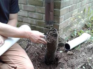 Person inspecting a clogged gutter downspout next to a brick wall.