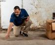 A man in a blue shirt kneels on the floor, conducting a home inspection in a dimly lit room with a cardboard box nearby.