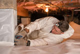 Man in protective suit installs a crawl space vapor barrier, securing plastic sheeting with tools while wearing a headlamp.