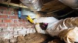 Person in protective gear installs insulation and a crawl space vapor barrier in a space with brick walls and exposed ducts.