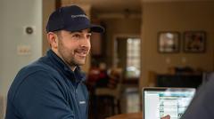 Man in a navy cap and jacket sitting at a table with a laptop, smiling and engaged in conversation.