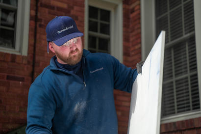 A man wearing a blue cap and sweatshirt works on a white board outside a brick building.