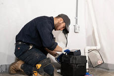 Person kneeling, inspecting a black sump pump in a basement with white walls and visible pipes.