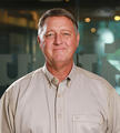 A smiling man in a beige shirt stands in a softly lit room with a blurred background.