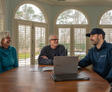 Three people sitting at a table with a laptop, having a conversation near large windows.