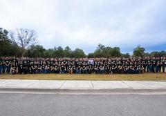 A large group of people wearing matching black shirts pose outdoors on grass, with trees and a cloudy sky in the background.