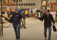 Workers in a warehouse, carrying tools and walking. Shelves with boxes and equipment in the background.