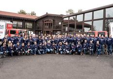 Large group of people in matching navy sweaters pose outside a log building with two red trucks nearby.