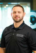 Man in a black polo shirt with "Foundation Recovery Systems" logo, standing and smiling in a bright room.