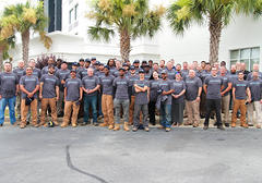 A large group of people wearing matching gray shirts poses together outdoors with palm trees in the background.