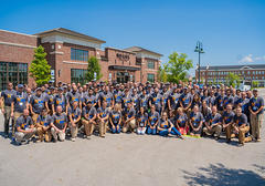 A large group of people in matching shirts posing in front of a brick building on a sunny day.