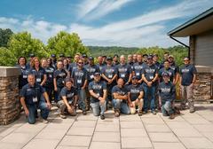 Group of people in matching shirts pose outdoors on a sunny day against a backdrop of trees and a blue sky.