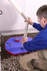 A person in a blue shirt expertly installs a PVC pipe onto a blue and red sump pump on gravel, ensuring optimal drainage for effective basement waterproofing.
