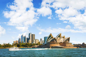 Sydney Opera House with city skyline in the background, viewed from the harbor under a partly cloudy sky.