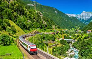 A train travels through a lush green mountain valley with blue skies and a river alongside.