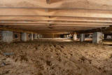 View of a crawl space under a house with exposed dirt ground and wooden floor joists above.