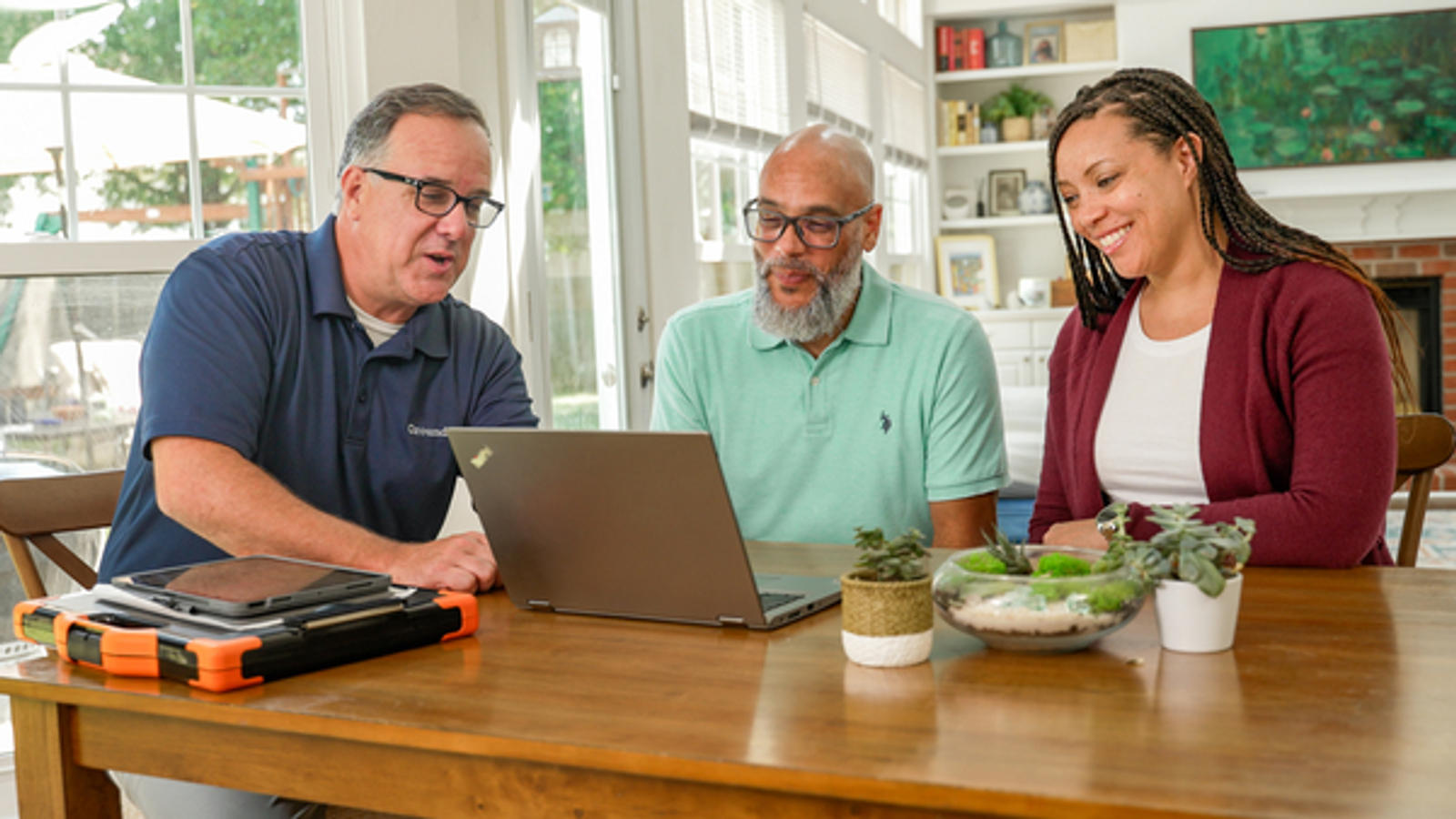 Three people sitting at a table, looking at a laptop screen, with indoor plants and a bookshelf in the background.
