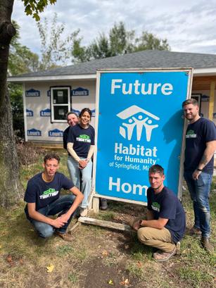 Volunteers in matching shirts pose by a "Future Habitat for Humanity Home" sign on a sunny day.