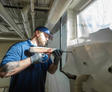 A man in gloves and a blue shirt uses a drill on a wall near a window in a basement.