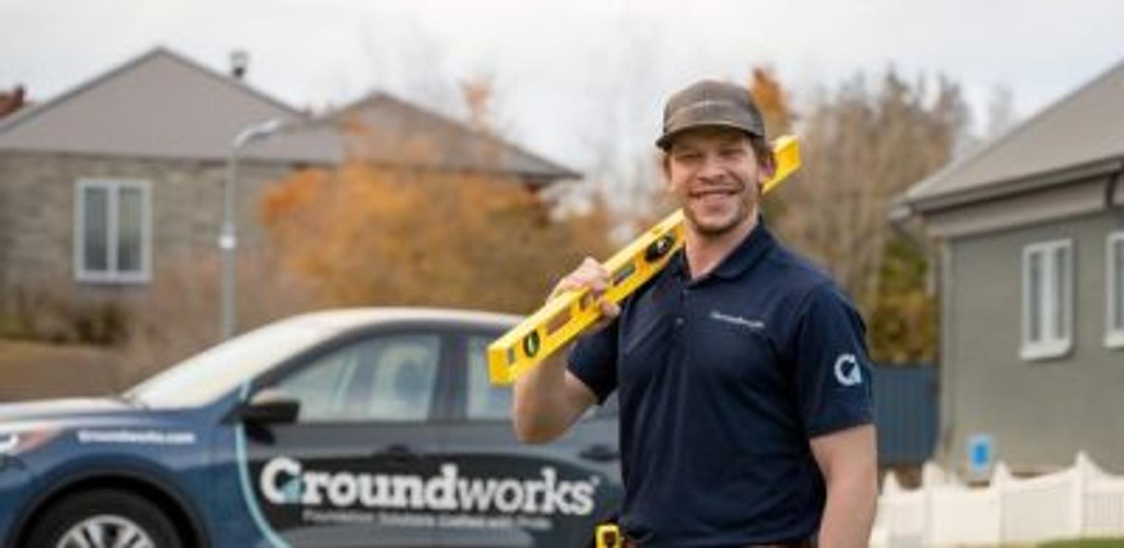 Smiling worker holds a level tool, standing outside near a Groundworks-branded car and houses.