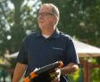 Smiling man in glasses holding a tablet outdoors, wearing a navy blue polo shirt.