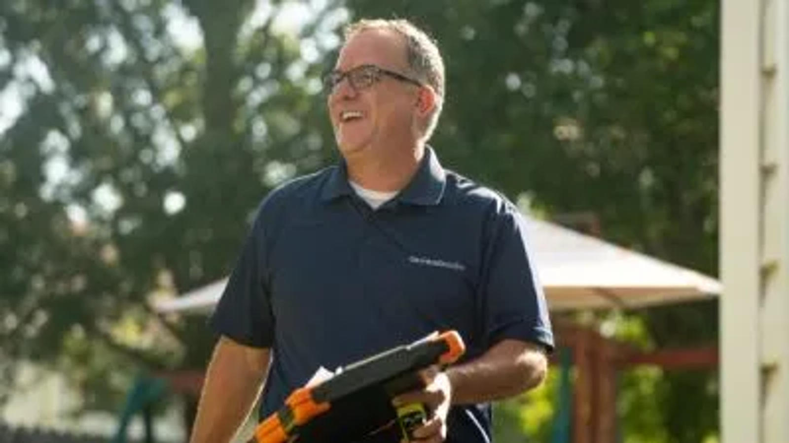Smiling man in glasses holding a tablet outdoors, wearing a navy blue polo shirt.