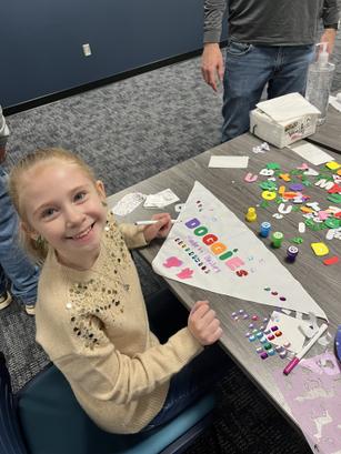 Smiling girl with a decorated banner at a craft table with letters and embellishments.