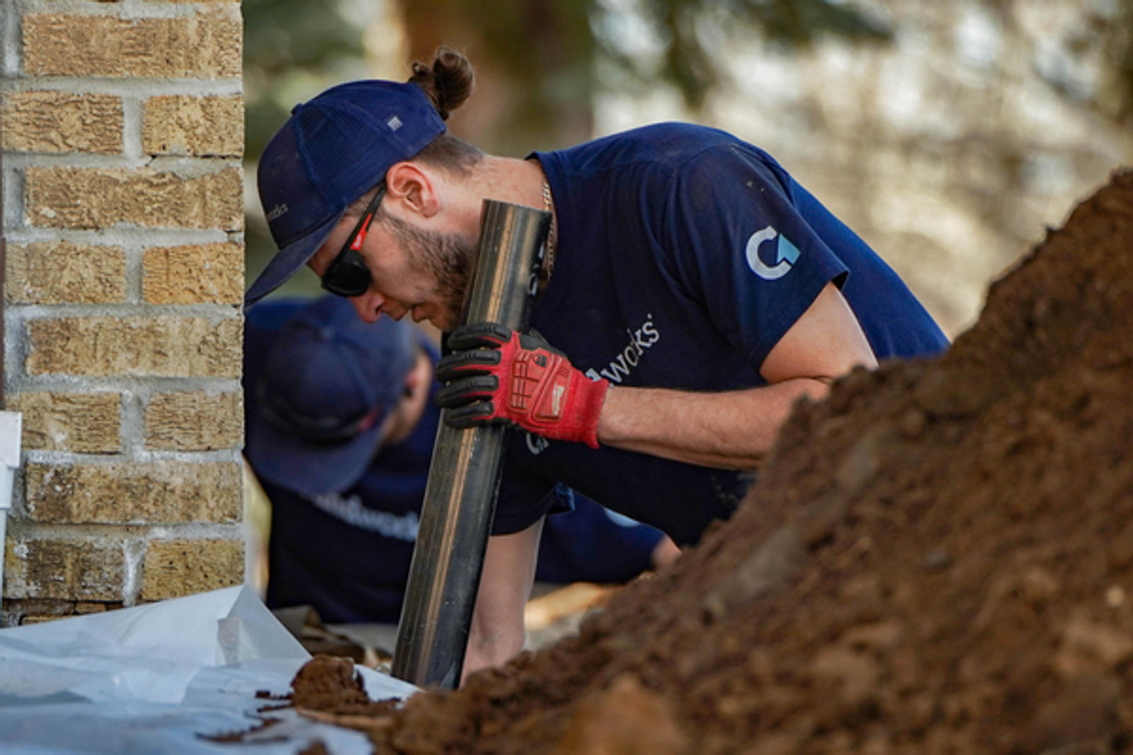 Person wearing a cap and gloves working on a construction site, holding a pipe near a brick wall.