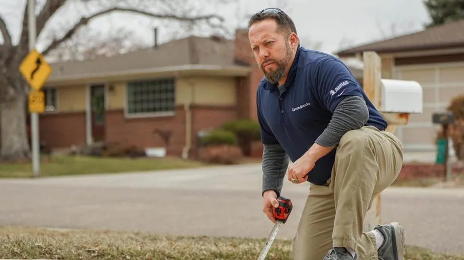 A man kneels on a lawn, using a tape measure, with a house and street in the background.