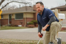 A man kneels on a lawn, using a tape measure, with a house and street in the background.