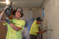 Two workers in bright shirts; one smiles holding a tool, the other installs pipes on a textured wall.