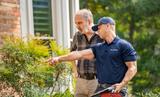 A man in a blue cap and uniform points, while another man listens in a garden setting.