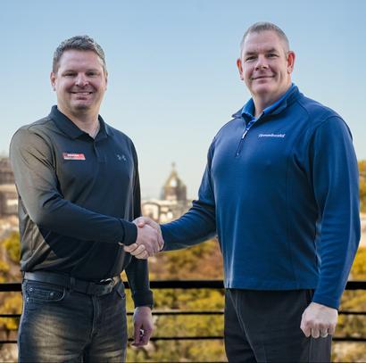 Two men smiling and shaking hands on a balcony with city buildings in the background.