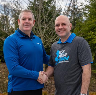 Two men shaking hands outside, dressed in blue and grey shirts, with trees in the background.