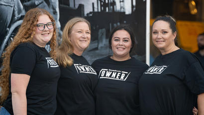 Four women in black "Owner" t-shirts smile and stand side by side in an outdoor setting.