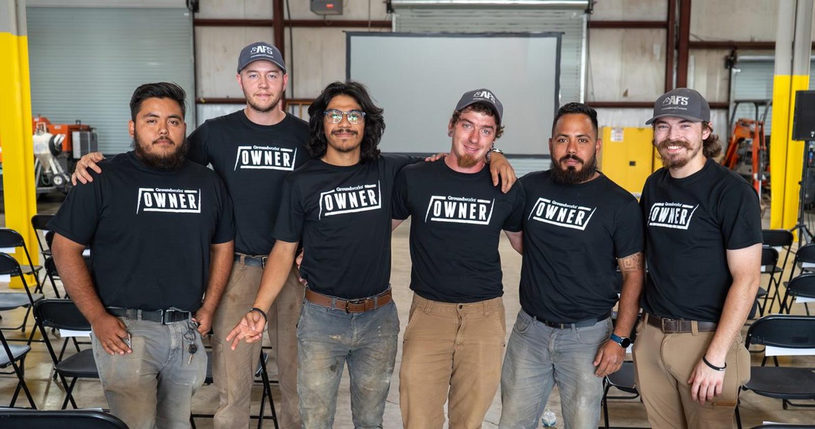 Six men wearing matching "Owner" t-shirts pose together in a workshop setting, smiling for the camera.