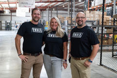 Three people in matching "Owner" shirts pose together in a warehouse with boxes and shelves in the background.