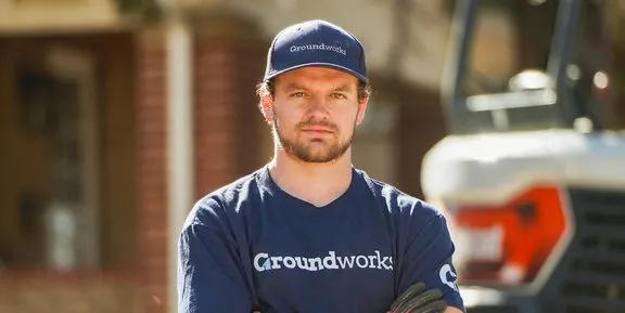 Man in a blue "Groundworks" shirt and cap stands with arms crossed in front of construction equipment.