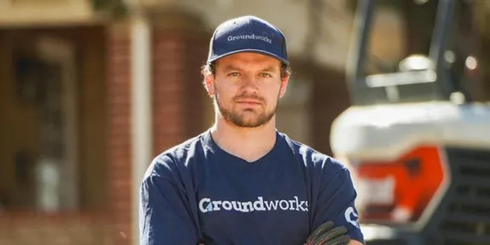 Man in a blue "Groundworks" shirt and cap stands with arms crossed in front of construction equipment.