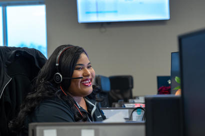 A woman with a headset smiles while working at a computer in an office.