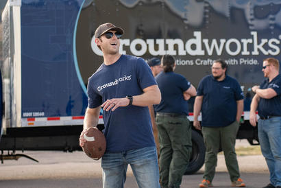 A man in a "groundworks" shirt throws a football with colleagues talking by a truck in the background.