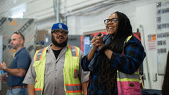 Two people smiling in a workshop, wearing safety vests and glasses.