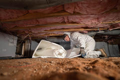Person in protective gear installing material in a crawl space with insulation and wiring visible above.