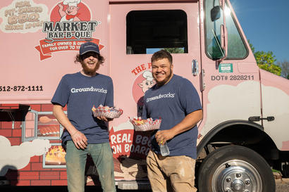Two men in blue shirts hold food trays in front of a pink food truck with "Market Bar-B-Que" branding.