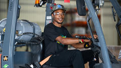 Person smiling while sitting on a forklift in a warehouse, wearing a cap and safety glasses.