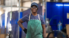 A smiling person in work attire stands confidently in an industrial setting.